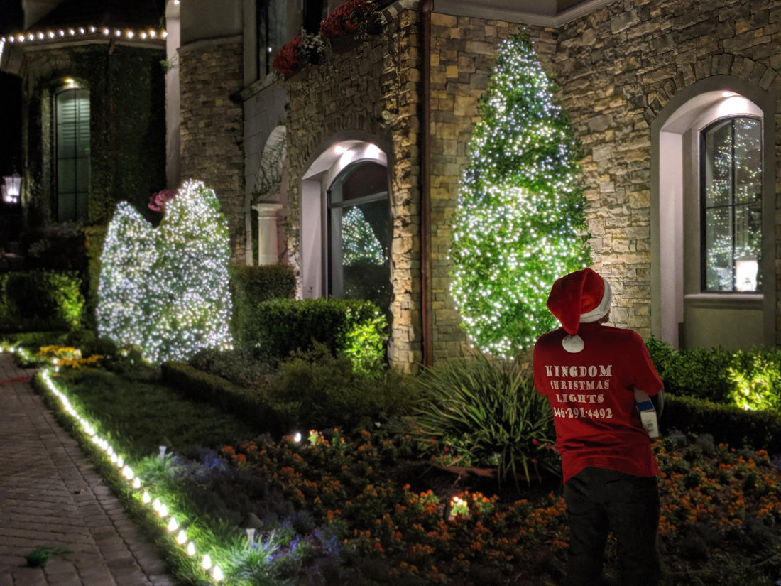 A Kingdom Christmas Lights team member in a Santa hat beside illuminated evergreens and a stone-front home during the 2019 Christmas season.