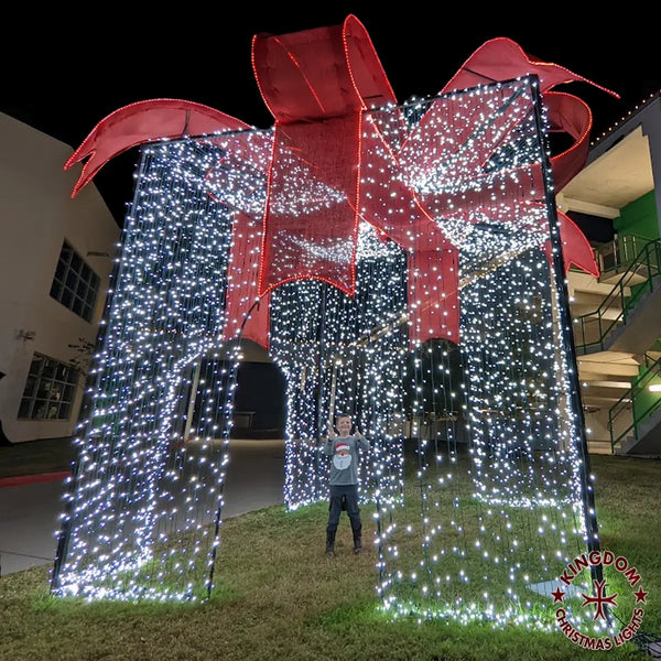 A large, walkable, presents-shaped Christmas decoration adorned with lights and a red bow, standing in a yard.