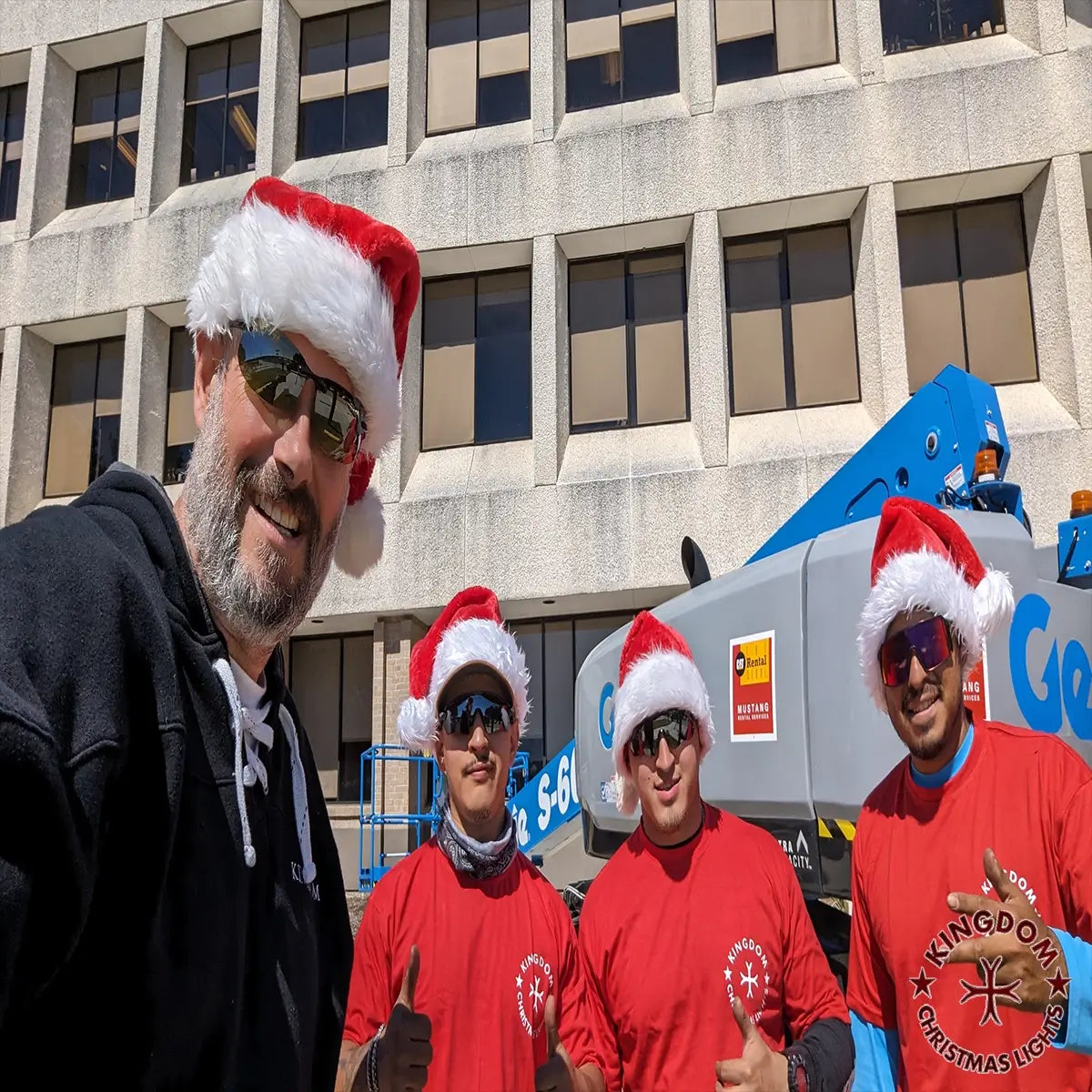 Four men wearing Santa hats and red shirts in front of a building with a crane.