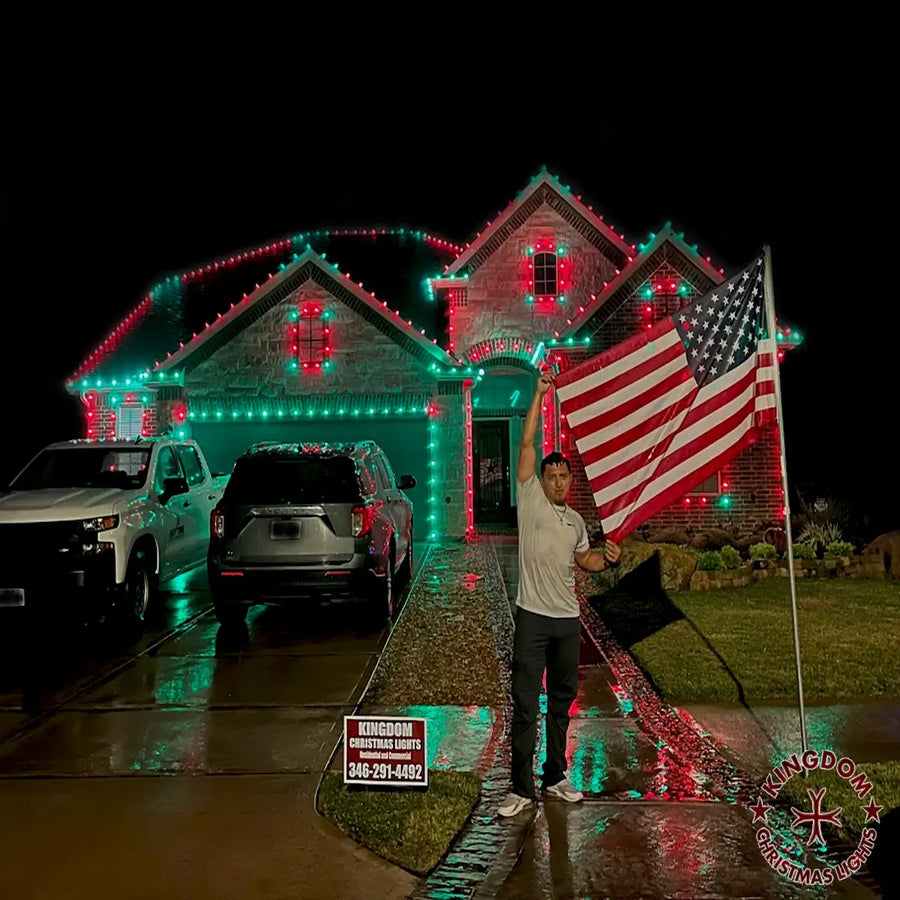 Person holding an American flag in front of a house decorated with Christmas lights