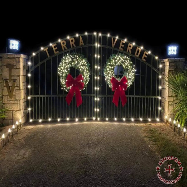 Decorative gate with wreaths and red bows at night, illuminated by string lights.