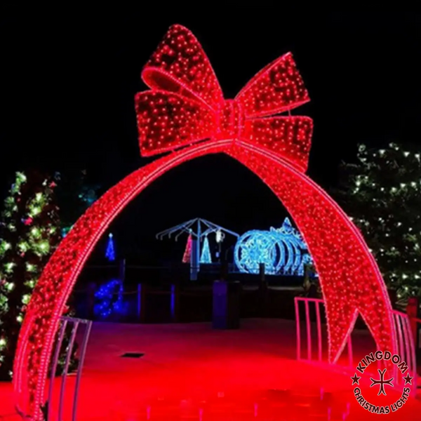 Decorative arch with a large red bow made of lights, set against a dark background with Christmas trees.
