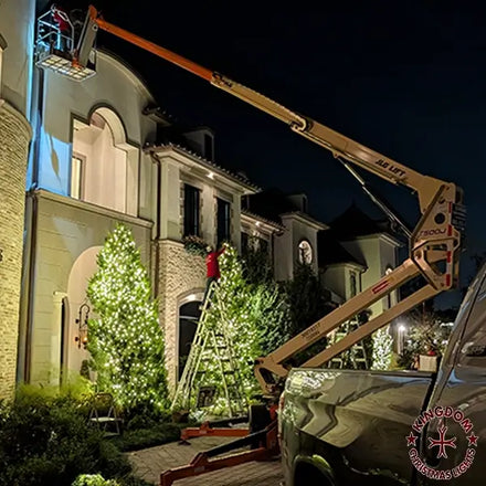 A towable lift being used to install Christmas lights on the exterior of a two-story house at night.